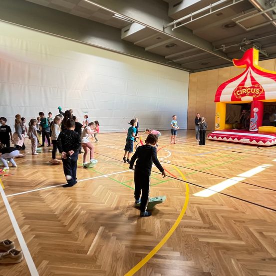 Eine Gruppe von Kindern spielt auf einem Holzboden, während ein großer aufblasbarer Zirkuszelt im Hintergrund steht. Einige werfen Bälle, während andere herumlaufen.