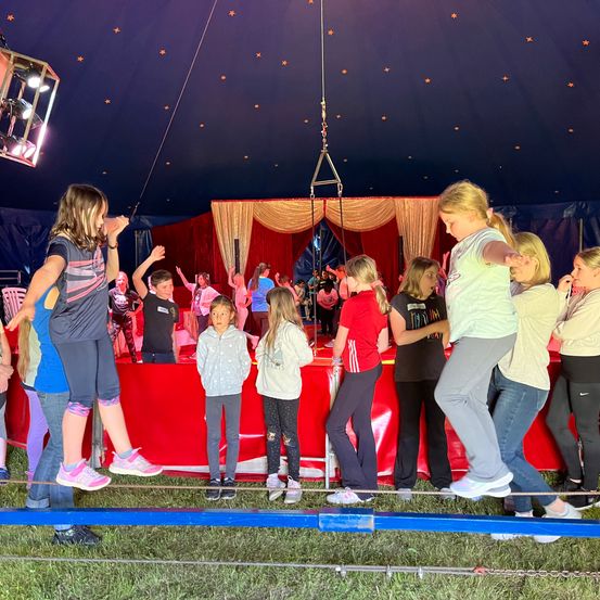 Mehrere Kinder balancieren auf einem Seil in einem Zirkuszelt, während andere zusehen. Im Hintergrund befindet sich eine rote Bühne mit rotem Vorhang.