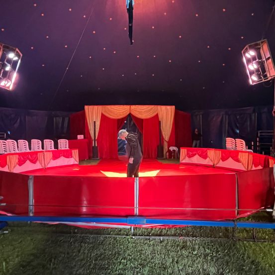 Ein Mann steht auf einer roten Plattform in einem Zirkuszelt, umgeben von Stühlen und Scheinwerfern.