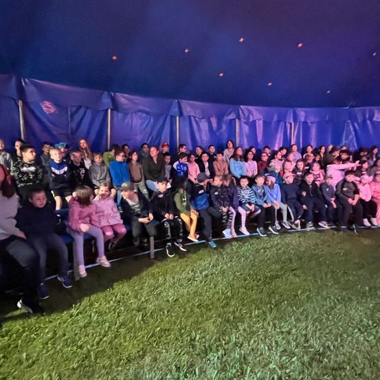 Eine große Gruppe von Menschen, einschließlich Kinder, versammelt sich unter einem blauen Zelt mit blauen Vorhängen und sieht sich eine Show an.