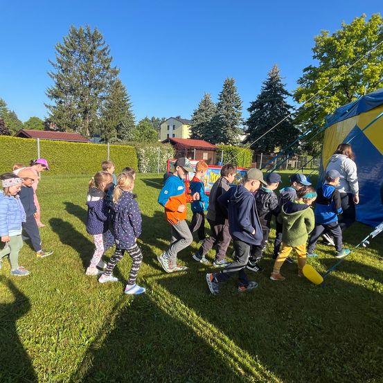 Eine Gruppe von Kindern läuft auf einem Rasenfeld mit einem Zirkuszelt im Hintergrund. Bäume und Büsche sind in der Nähe.