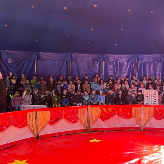 Eine Menschenmenge, einschließlich Kinder, sieht eine Zirkusaufführung in einem Zelt mit einem roten Boden und einer blauen Decke, die mit Sternen verziert ist.