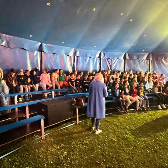 Eine Gruppe von Kindern und Erwachsenen sitzt unter einem blauen Zelt mit Sternen. Eine Frau in einem blauen Mantel steht vor ihnen.