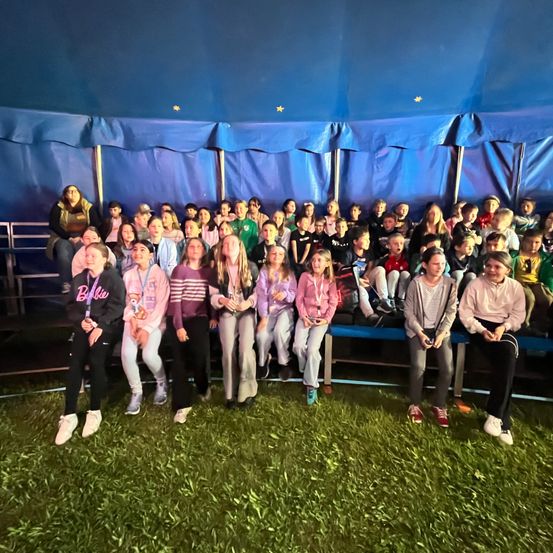 Eine Gruppe von Kindern und Erwachsenen sitzt unter einem blauen Zelt mit Sternmuster. Sie tragen verschiedene Kleidungsstücke und Schuhe. Einige stehen auf dem Grasboden.