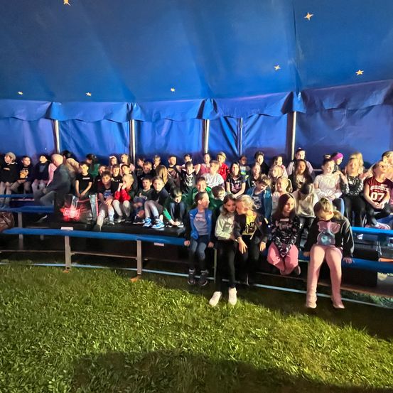 Eine Gruppe von Kindern sitzt auf Tribünen in einem blauen Zirkuszelt und blickt unter einem sternenbesetzten Dach nach vorne.
