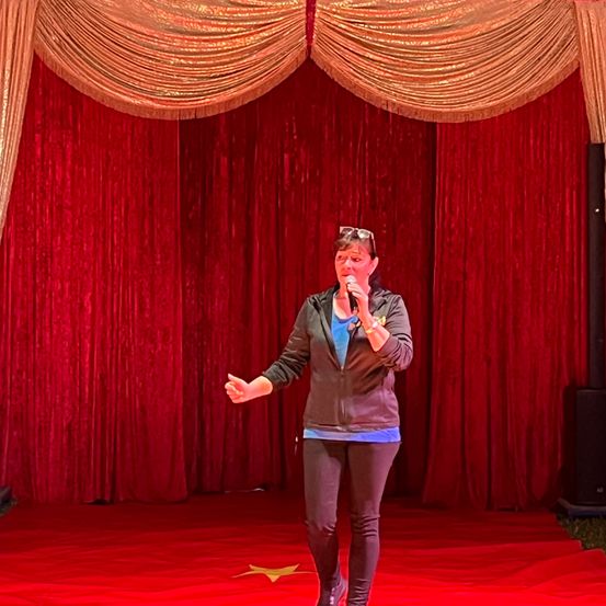Eine Frau in einer Jacke und Hose steht auf der Bühne und hält ein Mikrofon. Die Bühne hat einen roten Hintergrund und einen roten Boden mit einem gelben Stern.