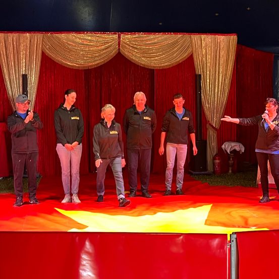 Eine Gruppe von Menschen in schwarzen Jacken und Turnschuhen steht auf einer roten Bühne, eine Person spricht in ein Mikrofon. Dahinter sind ein Feuerlöscher und ein Tisch mit einem weißen Tuch zu sehen.