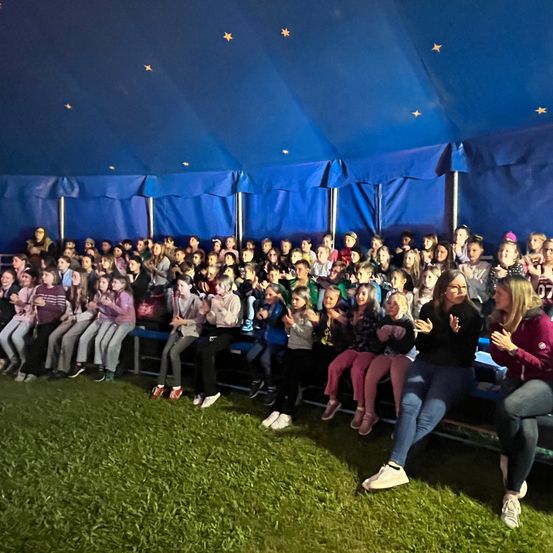 Eine Menschenmenge aus Kindern und Erwachsenen sitzt unter einem blauen Zelt, beleuchtet von Lichtern. Das Publikum ist engagiert, einige klatschen und andere halten ihre Hände hoch.