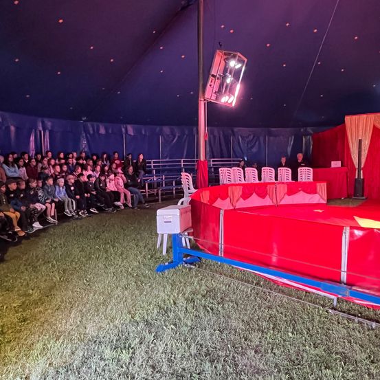 Ein Zirkuszelt mit einem roten Ring. Ein Publikum sitzt darum herum und schaut eine Vorstellung. Das Zelt ist mit roten und blauen Farben geschmückt.