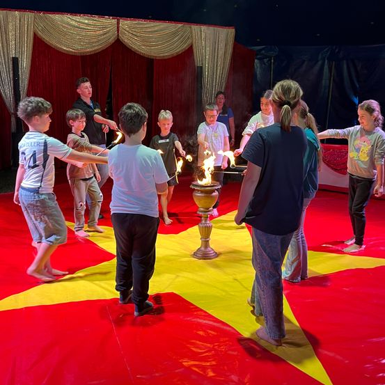 Eine Gruppe von Kindern und Erwachsenen führt einen Zirkusakt mit brennenden Fackeln auf einem roten und gelben Teppich auf. Ein Mann steht in der Mitte und hält eine Fackel, während andere brennende Fackeln halten.