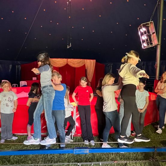 Eine Gruppe von Kindern balanciert auf einem Seil in einem Zirkuszelt. Sie werden von Erwachsenen unterstützt. Das Zelt ist mit roten Vorhängen und einem roten Tisch dekoriert.
