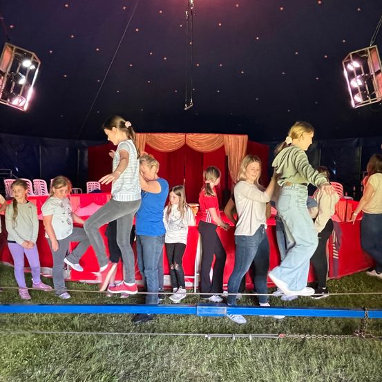 Mehrere junge Mädchen üben das Gehen auf einem Seil, unterstützt von Erwachsenen. Sie sind drinnen, in einer Zirkusumgebung mit roten Vorhängen und Beleuchtung.