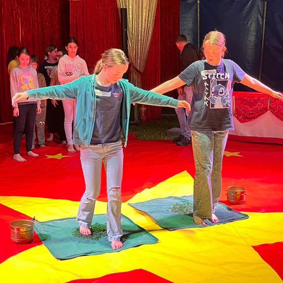Zwei Mädchen stehen barfuß auf grünen Tüchern mit Kieselsteinen in einem Zirkuszelt. Eines trägt eine blaue Jacke, das andere ein Grafik-T-Shirt. Zuschauer beobachten von hinten.