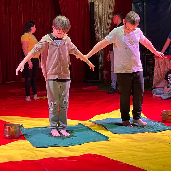 Zwei junge Jungen balancieren auf einem Zirkusboden mit rotem und gelbem Tuch. Sie sind barfuß und tragen eine Brille. Eine Frau in Gelb steht hinter ihnen.