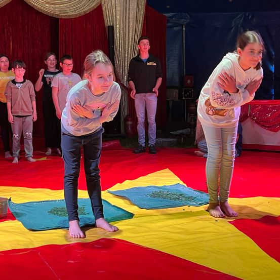 Mehrere Kinder stehen auf einer Zirkusbühne unter einem blauen Zelt. Zwei Mädchen vorne kauen auf einem gelb-roten sternförmigen Teppich.