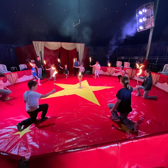 Eine Gruppe von Kindern und Erwachsenen führt einen Zirkusakt unter einem Zelt auf. Sie halten flammende Fackeln und knien auf einem roten Teppich mit einem gelben Stern.