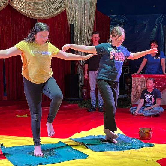 Zwei Frauen üben das Balancieren auf farbenfrohen Matten. Eine trägt ein gelbes Shirt, die andere ein blaues. Hinter ihnen beobachtet ein Mann. Im Hintergrund befinden sich ein roter Vorhang und ein Feuerlöscher.