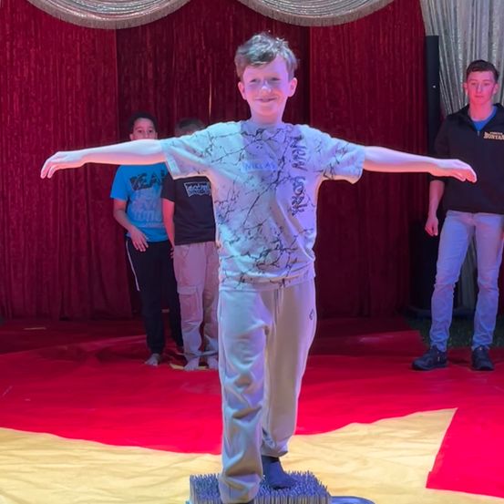 Ein junger Junge steht auf einem mit Nägeln versehenen Teppich, lächelt und posiert mit ausgestreckten Armen für ein Foto, während andere hinter ihm auf einem roten Teppich stehen.