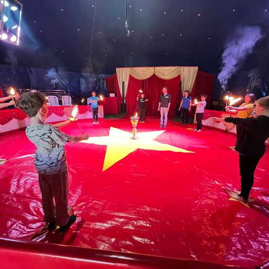 In einem Zirkuszelt stehen eine Gruppe von Kindern mit Fackeln um einen großen gelben Stern auf dem roten Boden. Ein Erwachsener steht in der Nähe eines goldenen Weihrauchbrenners.