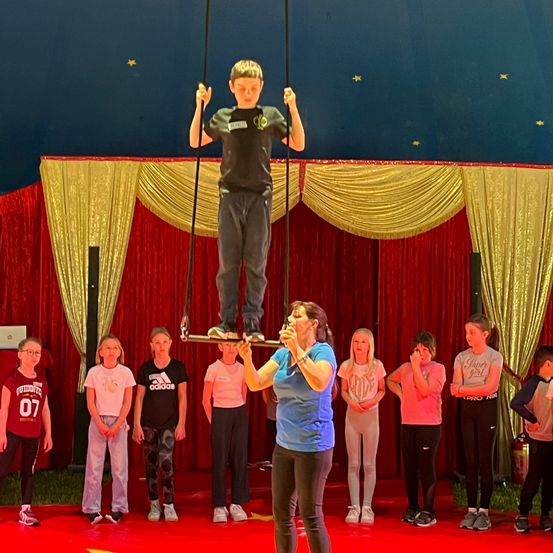 Ein junger Junge in einem schwarzen Shirt und Hosen führt eine Trapez-Vorführung unter einem blauen Zelt mit Sternen durch. Eine Frau hält das Trapez, während Kinder zusehen.