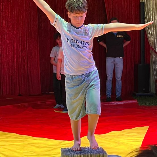 Ein Junge in einem weißen Fußballtrikot mit Emirates Fly Better steht auf einem mit Stacheln besetzten Teppich und balanciert. Hinter ihm beobachtet eine Person. Die Umgebung umfasst einen roten Vorhang und einen gelben und roten Boden.