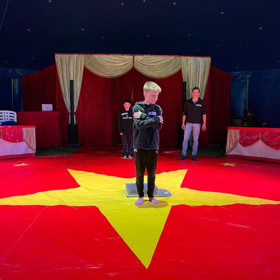 Ein junger Junge steht auf einem roten und gelben sternförmigen Teppich in einem Zirkus, mit zwei Erwachsenen hinter ihm. Die Szene ist drinnen mit einem roten Hintergrund und einer blauen Decke.