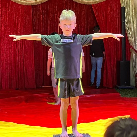 Ein junger Junge steht auf einem gelben und roten Teppich auf einer Bühne, Arme ausgestreckt, trägt ein schwarzes und gelbes Shirt. Im Hintergrund steht eine Person in der Nähe eines roten Vorhangs.