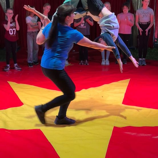 Ein Künstler hält ein Kind kopfüber, während eine andere Person von der Seite hilft. Zuschauer stehen hinter ihnen. Der Boden ist mit einem rot-gelben sternförmigen Tuch bedeckt.