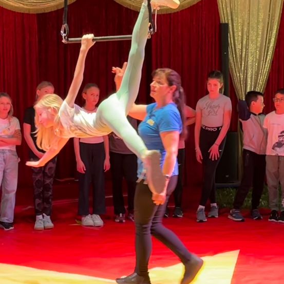 Ein junges Mädchen führt mit einer Trainerin einen Trapezakt in einem Zirkus aus. Zuschauer beobachten von hinten.