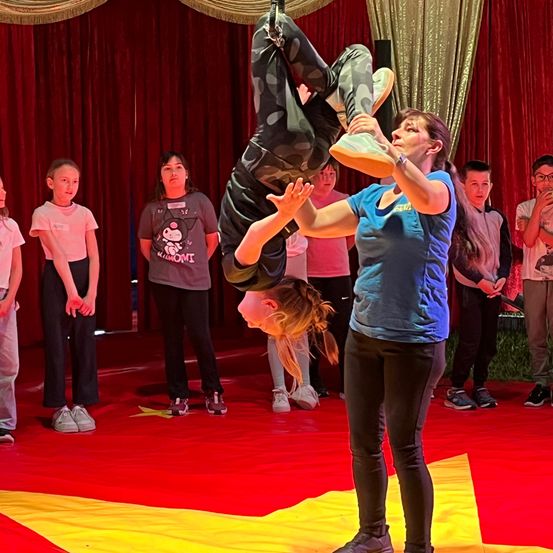 Ein junges Mädchen führt einen Handstand auf einer roten Bühne mit gelben Sternen aus. Eine Trainerin hält ihre Füße. Mehrere Kinder beobachten hinter ihnen auf einem roten Teppich stehend.