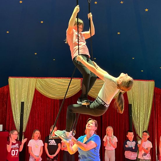 Zwei Frauen führen eine Trapez-Kunst in einem Zirkuszelt vor, wobei eine auf einer Stange balanciert und die andere die Vorführende hochhält. Zuschauer schauen von unten zu.