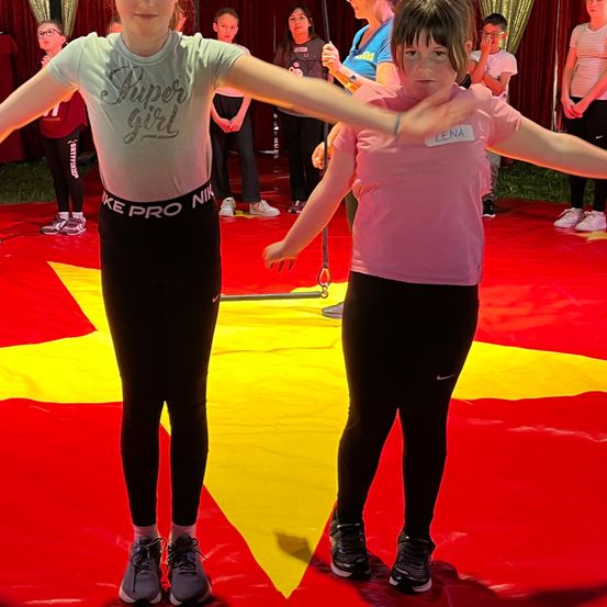 Zwei Mädchen stehen auf einem roten Teppich mit einem gelben Stern, beide in Sportkleidung. Eine trägt ein graues T-Shirt mit 'Super Girl' und 'Nike Pro'. Die andere hat ein rosa T-Shirt mit 'LENA'. Hinter ihnen stehen weitere Personen, einige mit Brillen.