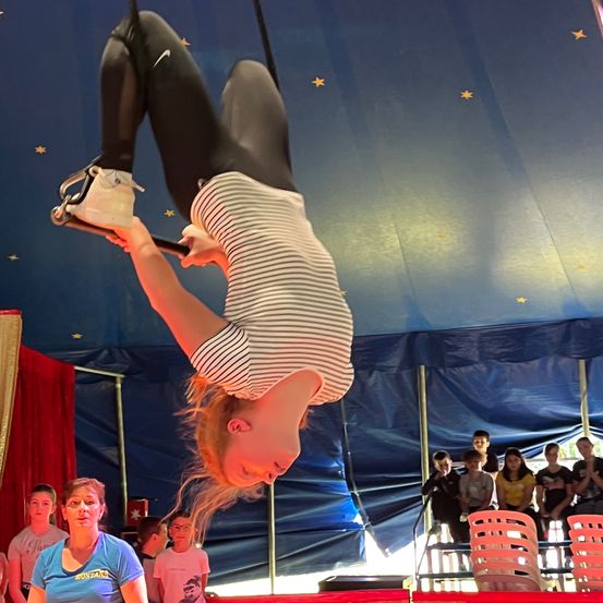 Eine Frau in einem gestreiften Shirt führt eine akrobatische Luftnummer in einem Zirkuszelt mit blauer Decke und gelben Sternen auf, während Zuschauer von den Stühlen unten zusehen.