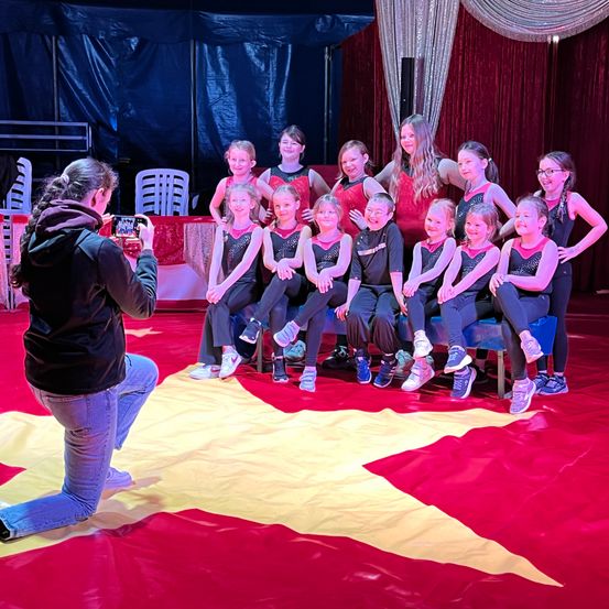 Eine Gruppe junger Mädchen in roten und schwarzen Outfits sitzt auf einem roten Teppich mit einem gelben Stern. Eine Person kniet vor ihnen und hält eine Kamera. Hinter ihnen ist ein blauer Vorhang.