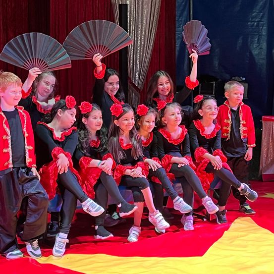 Eine Gruppe von Kindern in roten und schwarzen Kostümen posiert für ein Foto auf einer Bühne. Sie halten Fächer und sitzen auf einem rot-gelben Teppich.