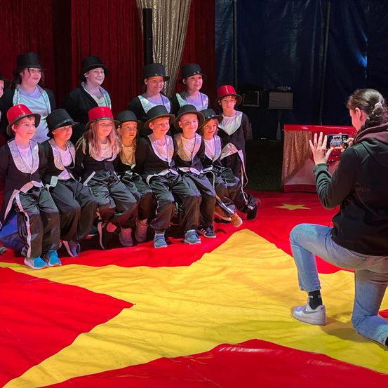 Eine Gruppe von Kindern in identischen Kostümen posiert für ein Foto in einem Zirkuszelt. Eine Frau kniet vor ihnen und macht ein Foto mit ihrem Handy.
