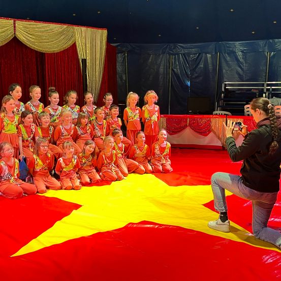 Eine Frau macht ein Foto von einer Gruppe von Kindern in passenden orangefarbenen Outfits, die in einem Kreis auf einem roten und gelben sternförmigen Teppich im Zirkuszelt sitzen.