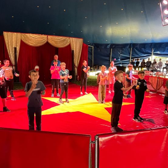 Kinder, die in einem Zirkuszelt auftreten, stehen auf einem roten Teppich mit einem gelben Stern in der Mitte. Das Publikum beobachtet sie hinter einer Barriere.