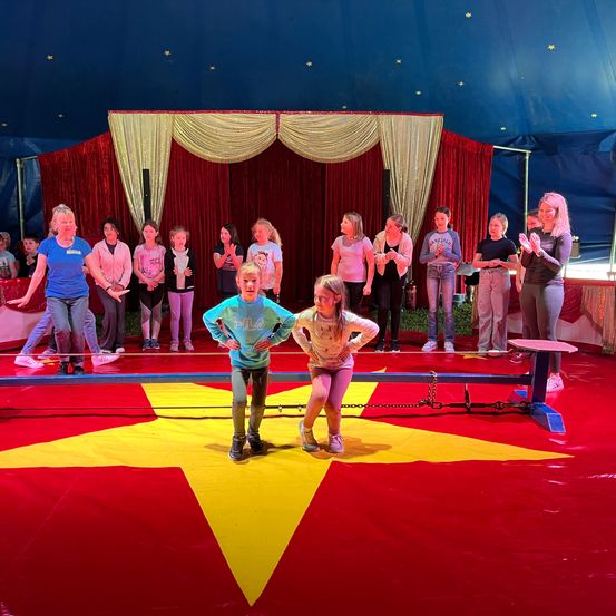 Eine Gruppe von Kindern und Erwachsenen versammelt sich in einem Zirkuszelt, wobei eine Frau und zwei Mädchen vor einem gelben Stern auf dem Boden stehen.