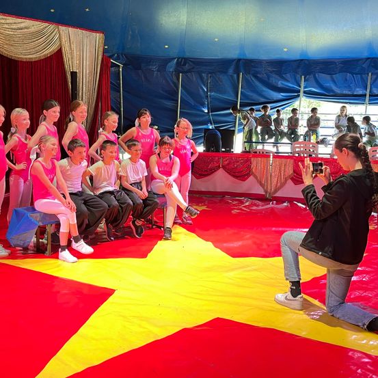 Eine Gruppe von Kindern in pinkfarbenen Uniformen sitzt auf einem roten und gelben sternförmigen Teppich in einem Zirkuszelt. Eine Frau in einer schwarzen Jacke macht ein Foto von ihnen mit einer Kamera.
