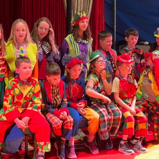 Eine Gruppe von Kindern in bunten Clown-Kostümen sitzt auf einer Bank in einem Zirkus. Einige tragen Hüte, Brillen und Schuhe. Hinter ihnen steht ein roter Vorhang.