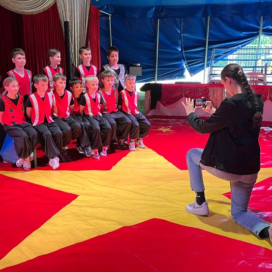 Eine Frau fotografiert eine Gruppe von Kindern in roten Westen, die auf einem roten Teppich mit einem gelben Stern in einem Zirkuszelt sitzen.