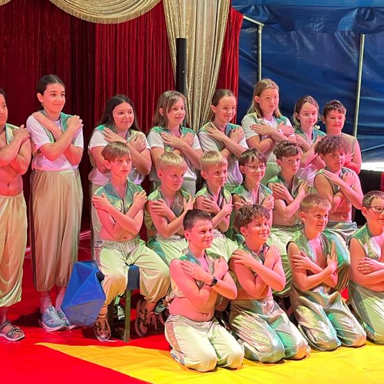 Eine Gruppe von Kindern in grünen Kostümen posiert für ein Foto auf einer Bühne, einige kniend und andere stehend. Ein roter und gelber Vorhang und ein blaues Zelt sind im Hintergrund.