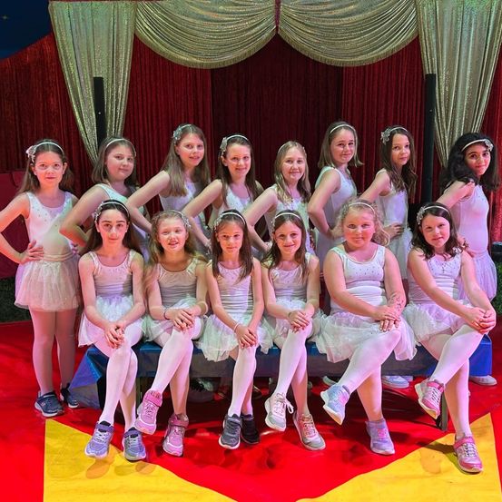 Eine Gruppe junger Mädchen in Ballettkostümen posiert für ein Bild auf einer Zirkusbühne mit rotem und gelbem Teppich.