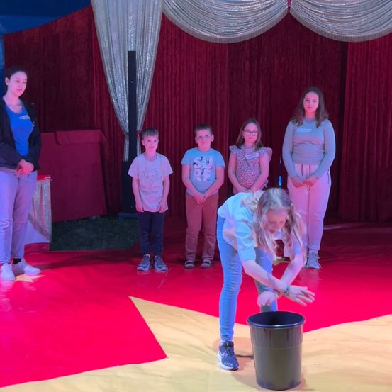 Eine Frau führt einen Zirkusakt auf der Bühne vor, während eine Gruppe von Kindern und Erwachsenen hinter ihr steht und zusieht.