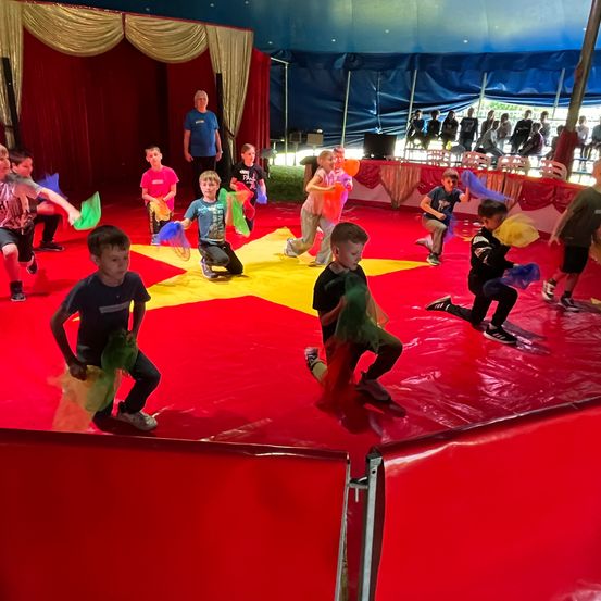 Kinder führen in einem Zirkuszelt mit einem roten und gelben Stern eine Darbietung auf und halten bunte Bänder. Ein Erwachsener beaufsichtigt. Zuschauer sitzen auf Bänken unter einem blauen Zelt.