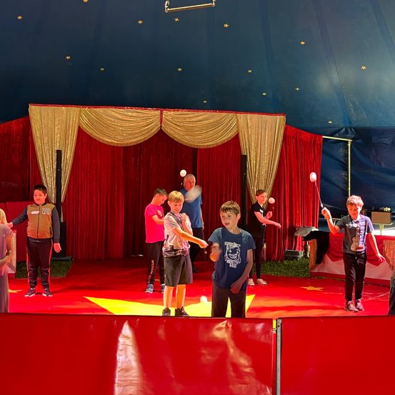 In einem Zirkuszelt führen mehrere Kinder einen Jonglierakt mit Bällen vor. Sie stehen auf einer roten Bühne, mit roten Vorhängen und goldenen Draperien im Hintergrund. Ein gelbes Sternmuster verziert den Boden.