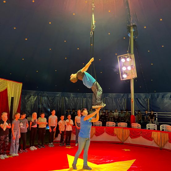 Ein Zirkusartist schwebt in der Luft, unterstützt von einem anderen Künstler am Boden. Das Publikum beobachtet die Szene von Stühlen und Stehplätzen aus unter einer sternförmigen Zeltdecke.