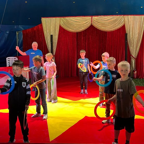 In einem Zirkuszelt halten mehrere Kinder und ein Erwachsener Hula-Hoops und führen einen Kreis auf einem roten und gelben Boden aus. Der Erwachsene gibt ihnen Anweisungen. Dahinter ein roter Vorhang mit goldenen Bordüren.