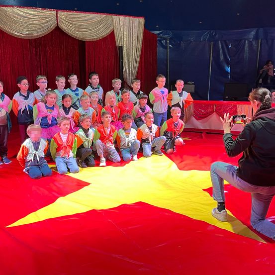 Eine Gruppe von Kindern in bunten Schals kniet auf einem roten Teppich mit einem gelben Stern in der Mitte. Ein Erwachsener kniet vor ihnen und macht ein Foto. Im Hintergrund befindet sich ein blaues Zelt und ein roter Vorhang.